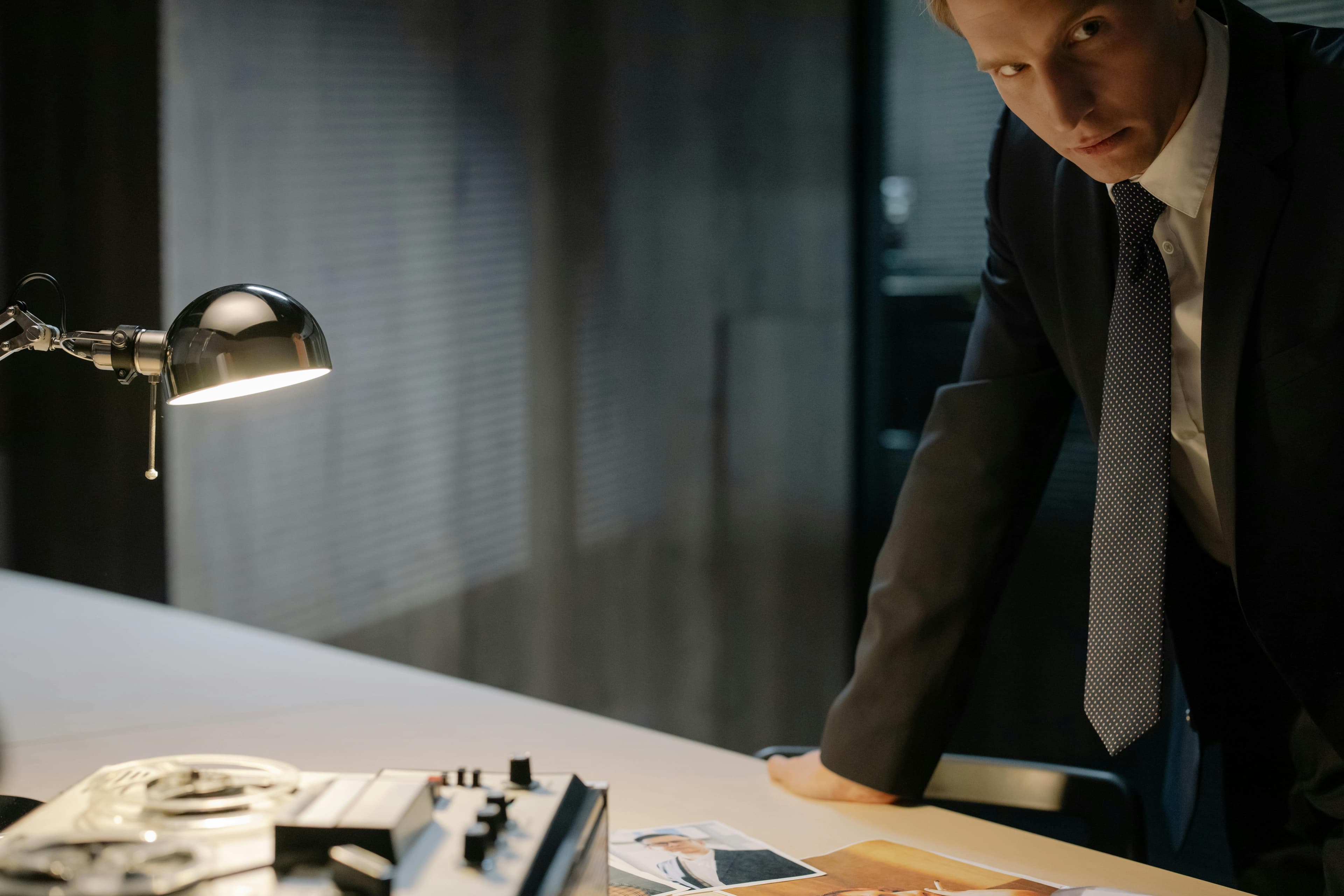 Man in a suit leaning over a desk with a lamp, photographs, and a tape recorder