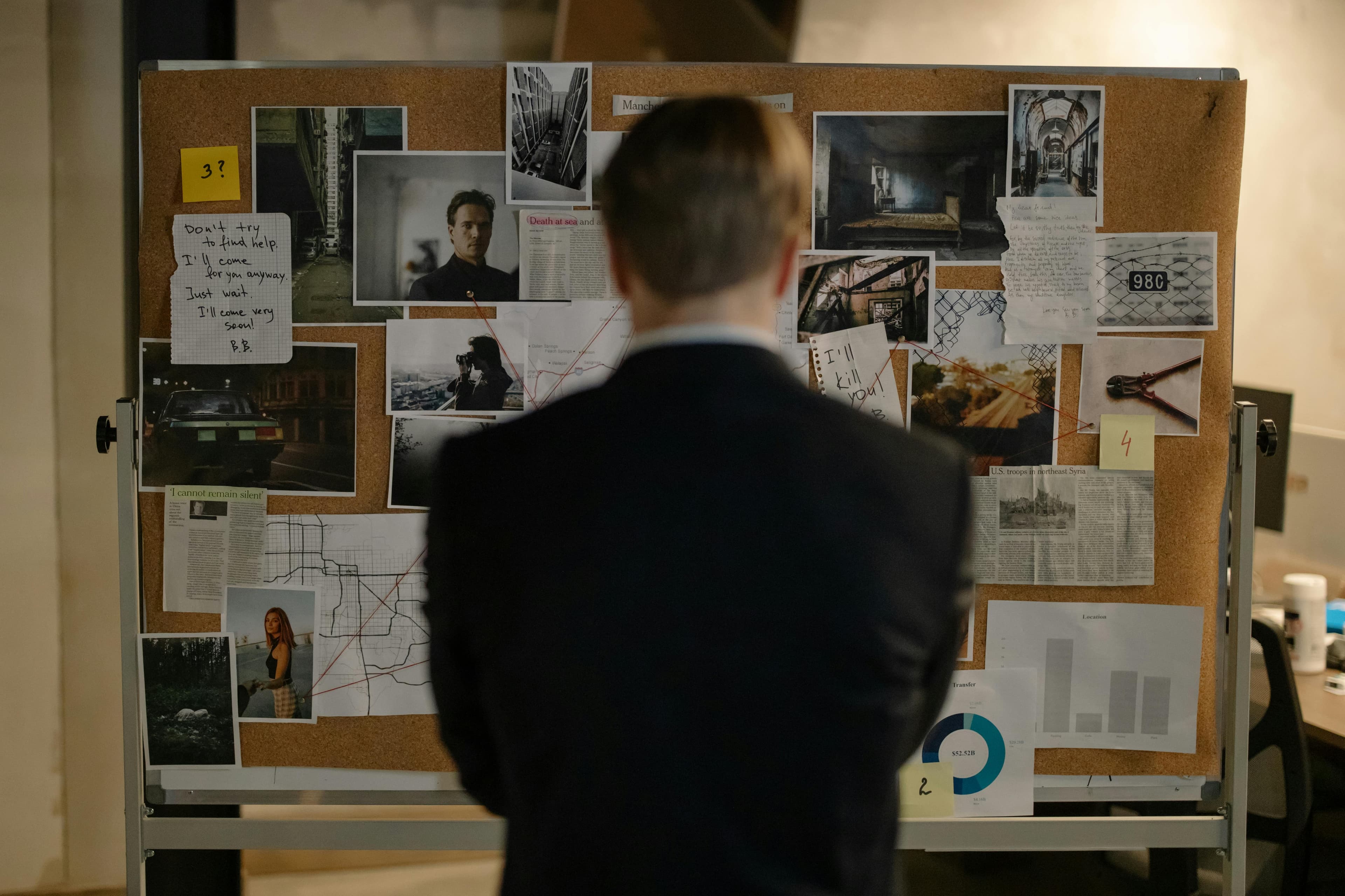 Man in a dark suit studying a case board of photos, notes, and a map